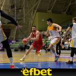 Basketball players in red and white uniforms compete for the ball on a indoor court, with spectators in the stands watching.